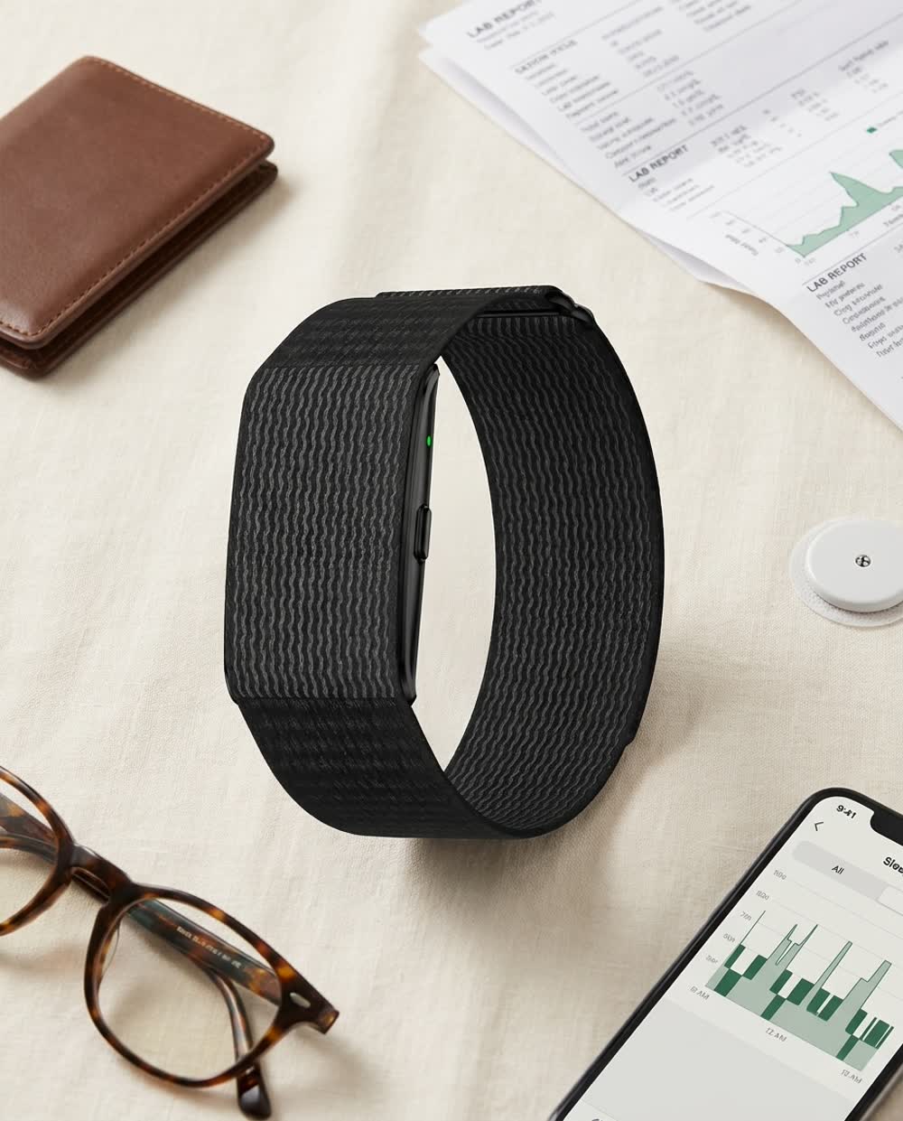 Pulsera Biometriv junto a una cartera, unos estudios de laboratorio, un sensor de glucosa y un teléfono con la app